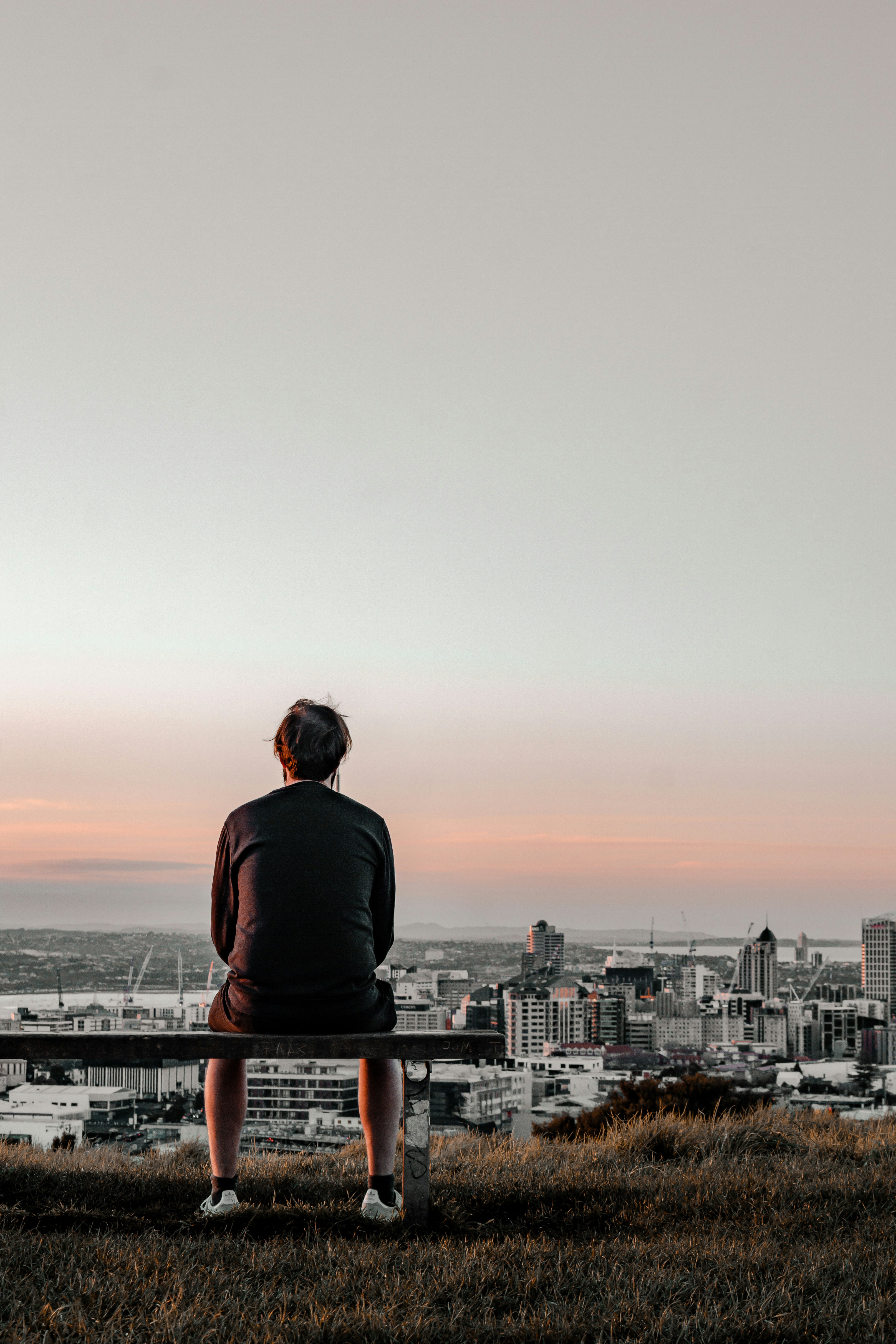 man in black long sleeve shirt sitting on brown wooden bench looking at city during daytime